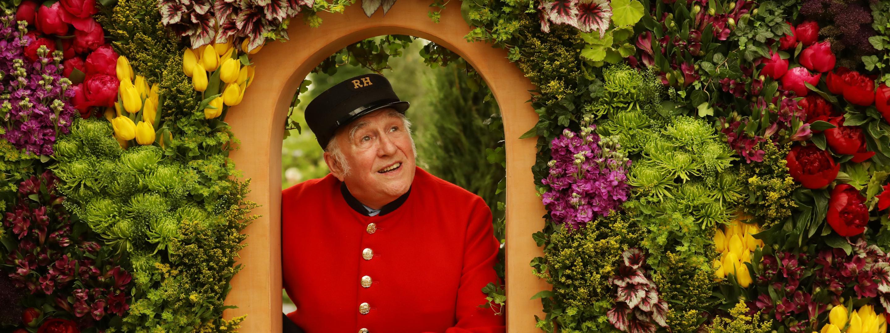 Pensioner in red and black RH uniform looking through gate surrounded by flowers