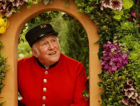 Pensioner in red and black RH uniform looking through gate surrounded by flowers