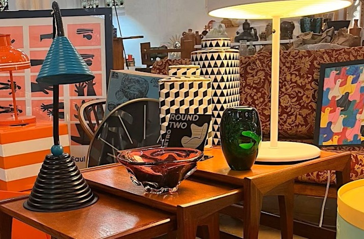 A display of colourful vintage lamps, glassware and patterned decor arranged on wooden tables at an indoor market.