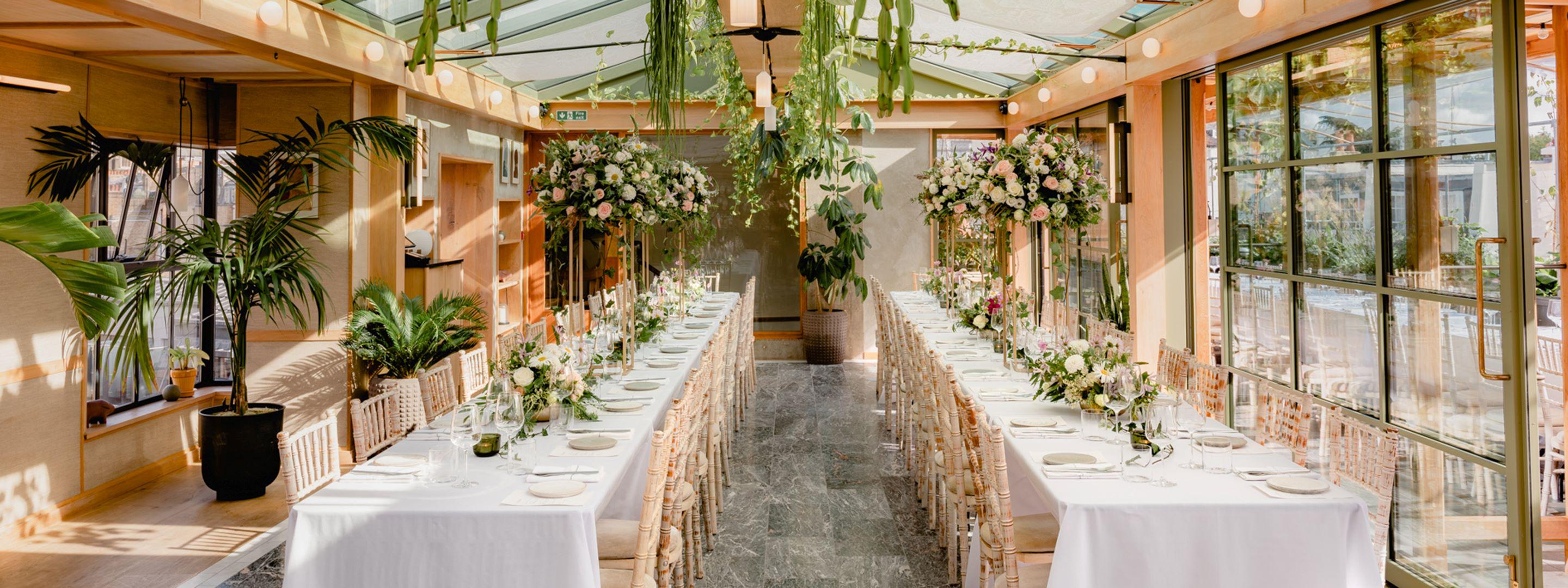 Large room with two long tables set with cutlery, plates, and flowers set up for a celebration.