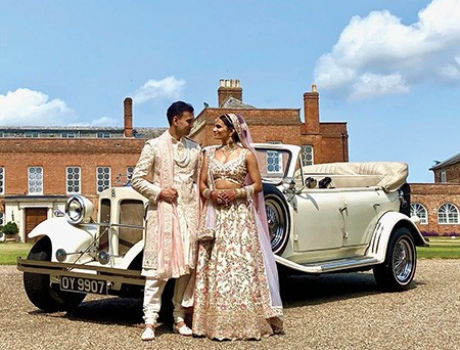 An Asian couple wearing a lehenga stand in front of a classic car next to a stately home in the English countryside