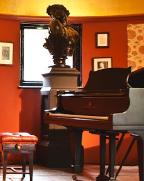 A piano with an bust sculpture in the background and paintings hanging on brown walls