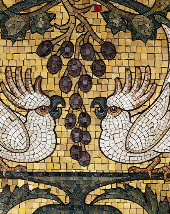 A close-up of a mosaic shows two white birds facing each other on a branch beneath a cluster of grapes, surrounded by ornate scrolling patterns