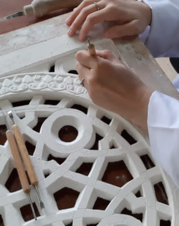 Hands use carving tools to shape an intricate lattice design on a large decorative panel.