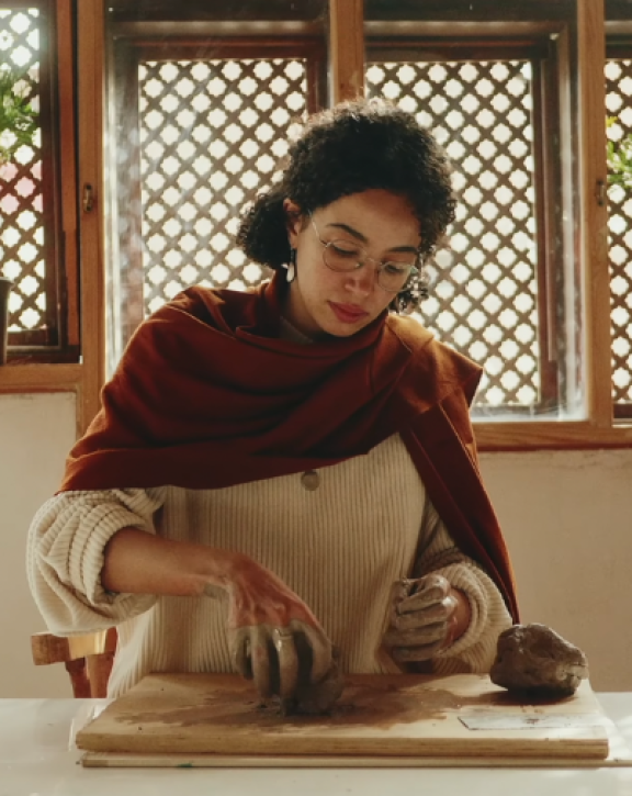 A person works with clay on a table in a sunlit room with lattice windows and potted plants in the background.