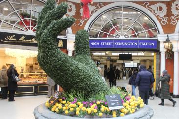 Large topiary sculpture shaped like an open hand, surrounded by yellow and purple flowers in a circular stone planter. The display is located inside High Street Kensington Station, with the station sign visible above the entrance in the background. Decorative shopfronts and people walking through the station are also visible.