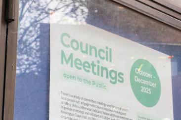 Poster displayed on a glass window announcing “Council Meetings open to the Public” in large green text, with a green oval stating “October to December 2025.” Additional smaller text below explains details about committee meetings and public participation.