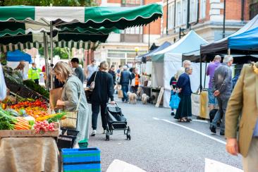 People shopping and walking through a busy outdoor street market lined with stalls selling fruit, vegetables and other goods