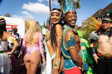 A black man and woman standing back to back in the middle of a street. Both are wearing carnival costumes.