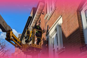 Person standing in a raised access platform carrying out maintenance work on the exterior of a multi-storey brick residential building, with windows and balconies visible above