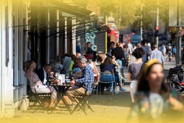 People sit at outdoor café tables along a busy high street, with pedestrians, shopfronts and bicycles visible along the pavement in an urban setting