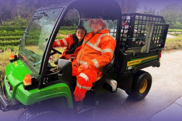 Alt text:  Two council workers wearing high-visibility orange workwear sit in a green utility vehicle on a roadway, with trees and greenery visible in the background