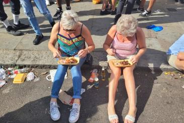 Two middle-aged women sitting on a curb eating street food during Notting Hill Carnival
