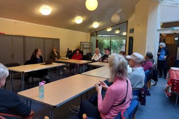 A group of people sit in a community meeting room arranged with tables in a U‑shape