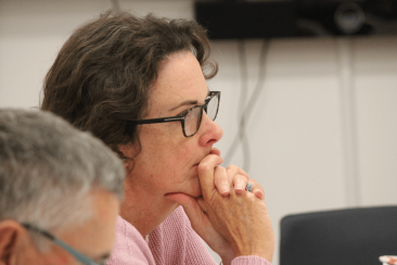 Close up of a person at a meeting with dark brown hair and dark brown rimmed glasses. They have their hand to their mouth in concentration and is wearing a ring. In the foreground is a softly blurred image of a man with grey hair and blue and black rimmed glasses looking down. 
