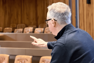 Person with short grey hair and glasses, seen from behind, gesturing with one hand while seated in a wood-panelled room with rows of empty brown chairs.