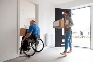 A man in a wheelchair and a woman behind him both enter a room carrying cardboard boxes