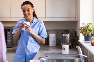 A woman in a nurses' outfit stands in a kitchen. She is leaning against a counter holding a mug.