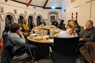 Group of people seated around a large table in a meeting room, with papers, drinks, and food containers on the table