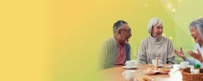 Three older adults sitting at a table, smiling and talking over tea and biscuits