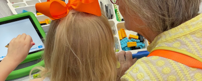 A young child plays on a tablet with an older person supervising