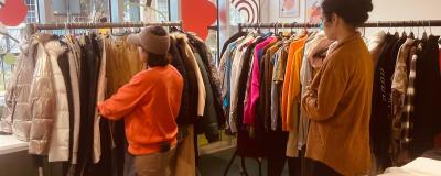 Two people browse a rack of winter coats and clothing inside a bright shop with large windows and tables of folded garments