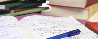 Open notebook with handwritten maths equations, a blue pen resting on the page, and stacked books in the background.