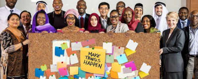 People gather around a cork board with post-it notes on it, with the middle one reading "Make Things Happen"