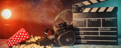 Stock photo of popcorn, a super eight camera, a clapperboard and a film cannister on a wooden floor with a yellow-red backlight on the far left.