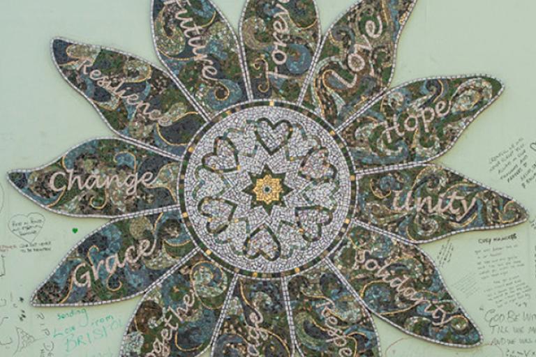 A photo of a mosaic in the shape of a flower hung on a light green wall. The centre of the flower has a gold star with six hearts around it. There are twelve petals surrounding the centre of the flower and each petal has a word on each petal. The words on the petals are “Forever, Love, Hope, Unity, Solidarity, Respect, Courage, Together, Grace, Change, Resilience, Future. 