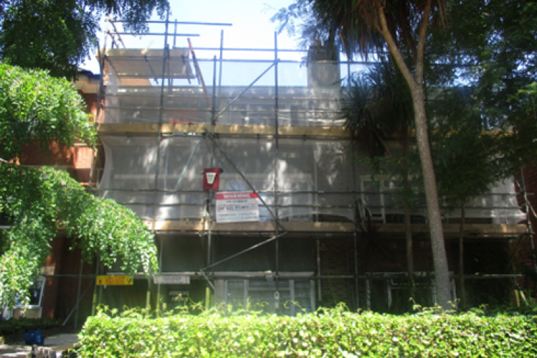 An image of a street property surrounding by trees either side and a hedge in front covered in scaffolding