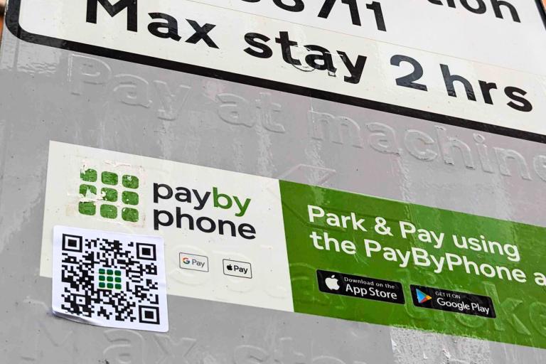 A parking sign showing legitimate pay by phone information and a sticker with a fake QR code