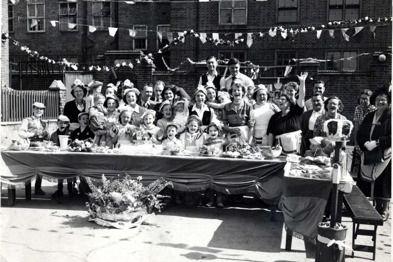 Black and white image shows a long table with lots of smiling and laughing people arranged behind it waving flags and celebrating the coronation