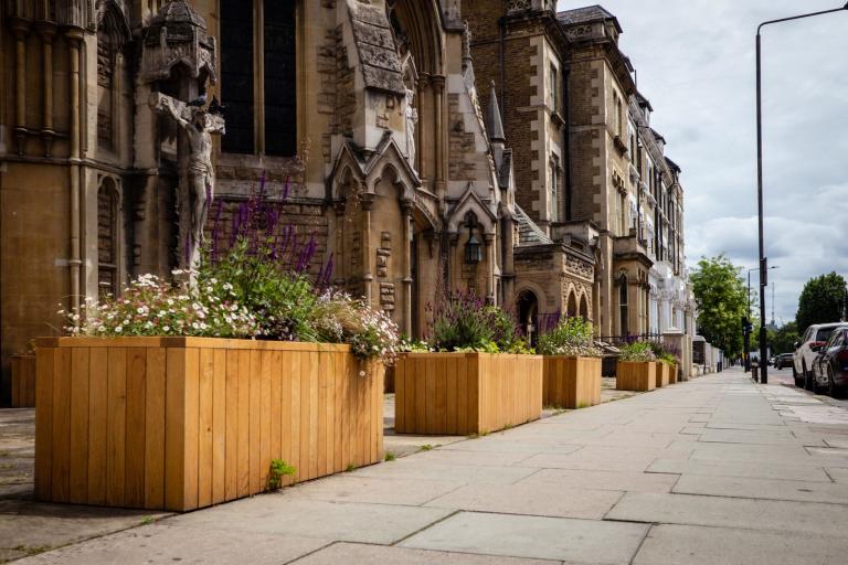 A pavement view of wooden box planters containing green, yellow and purple flowers in front of St John the Baptist Church