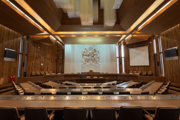 Large council chamber with tiered seating arranged in a horseshoe shape, wooden panelled walls, and a high ceiling featuring modern light fixtures.