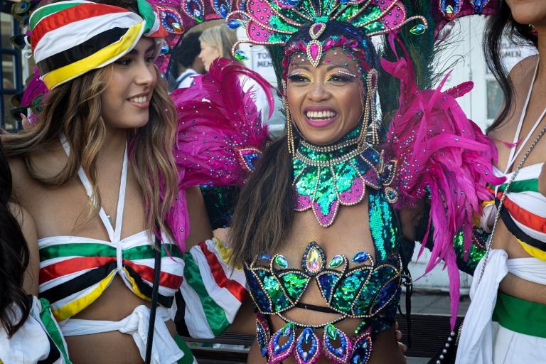 A woman centre-right dressed in turquoise and purple costume smiles just to the left of camera while a woman on left looks to her left wearing a red, white, black and yellow striped costume
