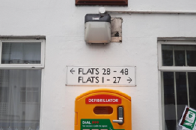 A bright yellow wall mounted defibrillator on the external wall of a block of flats