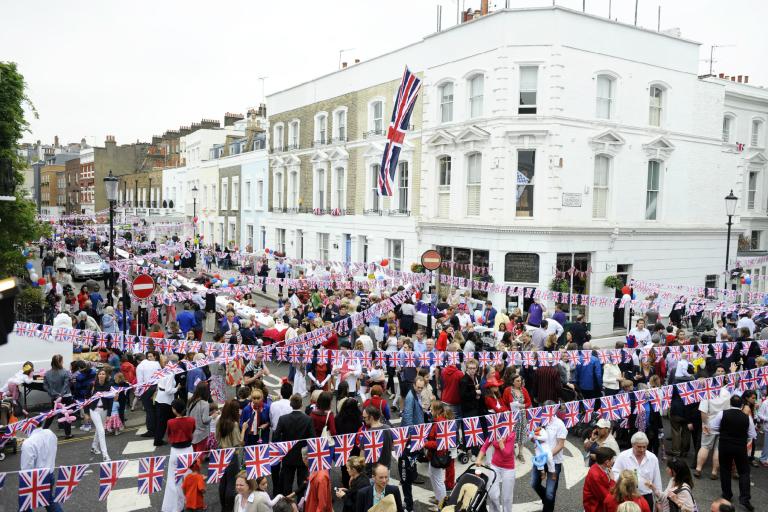 Image taken across intersection of two streets criss-crossed with Union Flag bunting and full of people celebrating with stone buildings above them 