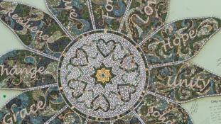 A photo of a mosaic in the shape of a flower hung on a light green wall. The centre of the flower has a gold star with six hearts around it. There are twelve petals surrounding the centre of the flower and each petal has a word on each petal. The words on the petals are “Forever, Love, Hope, Unity, Solidarity, Respect, Courage, Together, Grace, Change, Resilience, Future.