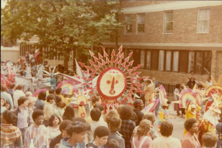 Colour photograph of a crowded street scene at Notting Hill Carnival, showing people walking past large, brightly decorated costume structures