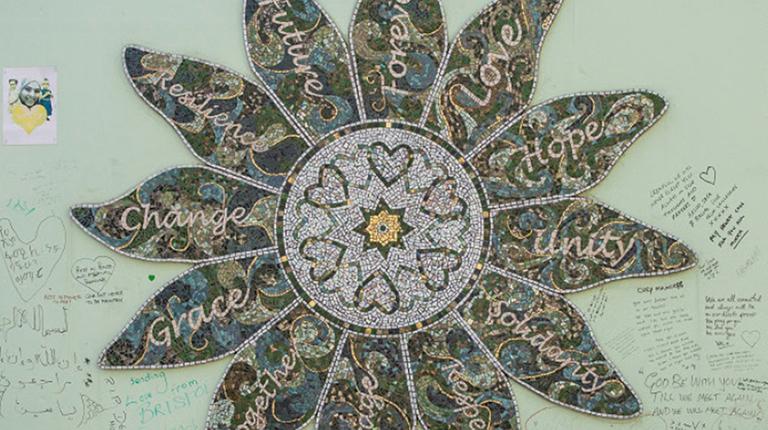 A photo of a mosaic in the shape of a flower hung on a light green wall. The centre of the flower has a gold star with six hearts around it. There are twelve petals surrounding the centre of the flower and each petal has a word on each petal. The words on the petals are “Forever, Love, Hope, Unity, Solidarity, Respect, Courage, Together, Grace, Change, Resilience, Future. 