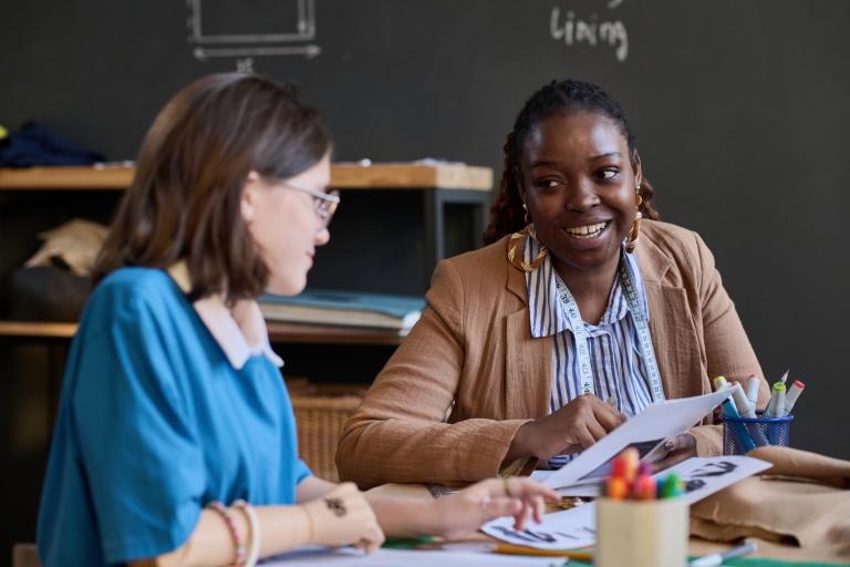 Two people sit at a table reviewing printed documents together, with stationery and materials spread out in a workshop or classroom setting