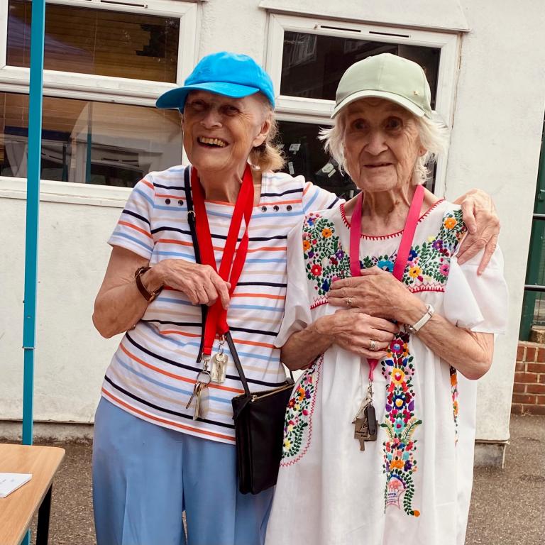 Image of two elderly ladies hugging and smiling wearing sun hats and summer dresses