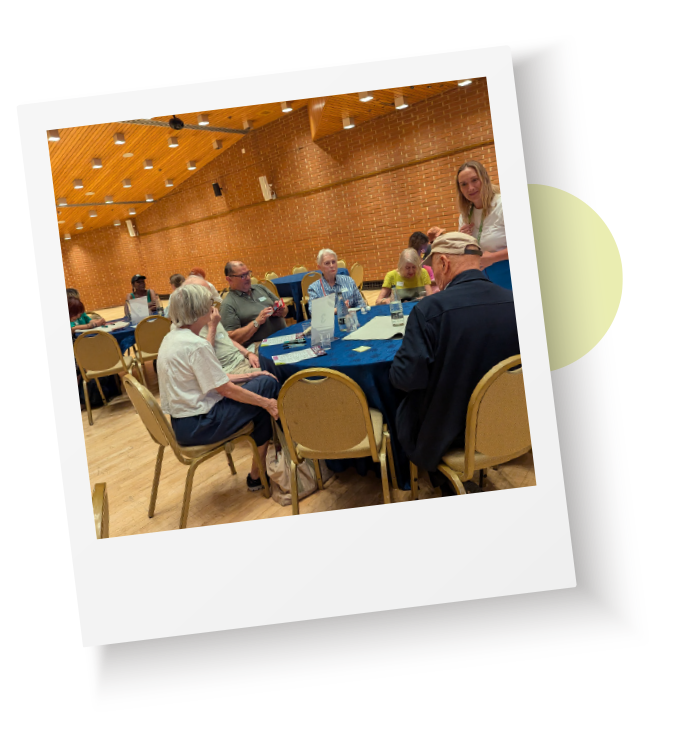 Residents taking part in a community meeting, seated around tables in a hall and discussing local issues and services