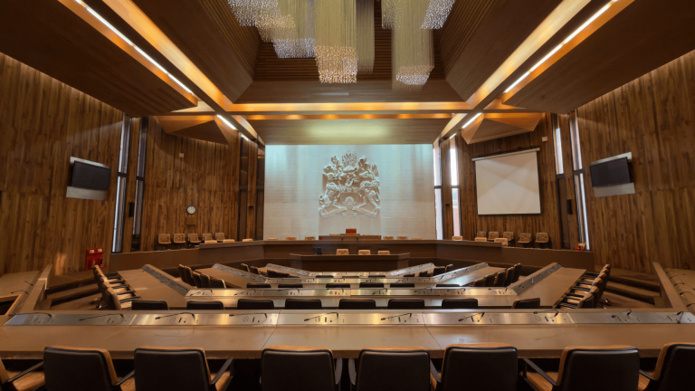 Large council chamber with tiered seating arranged in a horseshoe shape, wooden panelled walls, and a high ceiling featuring modern light fixtures.