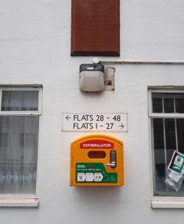 A bright yellow wall mounted defibrillator on the external wall of a block of flats