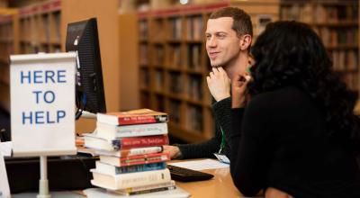 A male member of libraries staff sitting with a woman at a computer helping her to find the information she is looking for.