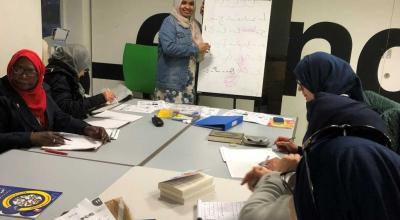 A group of Asian women sitting on a large bank of desks discussing ideas with one woman standing at the end of the desk writing the ideas on a flip-chart