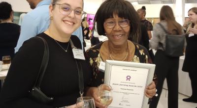 Two women stand smiling into the camera, lady on right holds up a framed white certificant