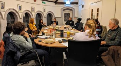 Group of people seated around a large table in a meeting room, with papers, drinks, and food containers on the table